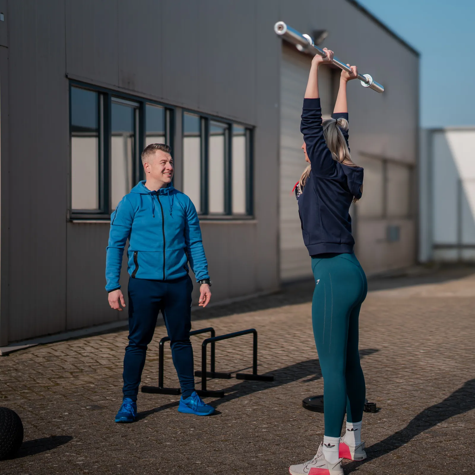 Arnold begeleidt buiten een training: een vrouw drukt een halterstang boven haar hoofd
